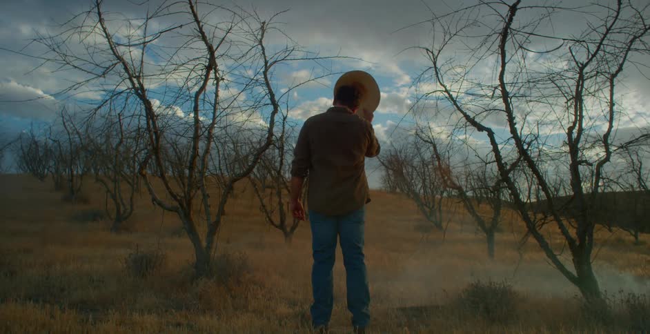 Cowboy taking off his hat while standing in a field with dry trees
