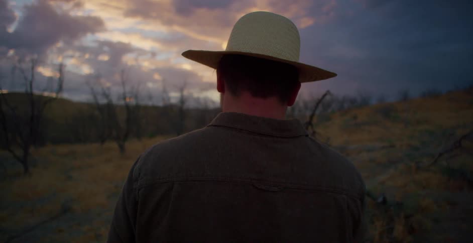A white man walks on a grassy plain and removes his hat to take in the scenery