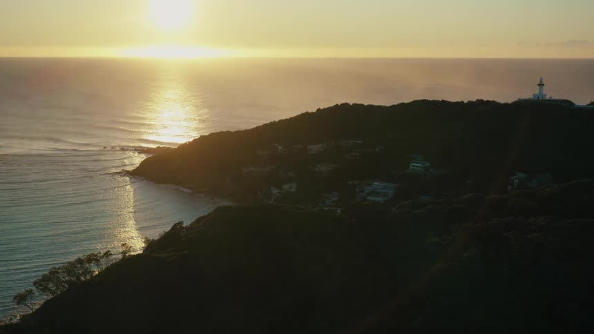 A lighthouse sits on a coastal rural area as waves wash ashore with the sun low in the sky