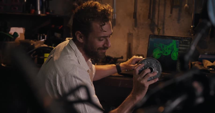 A mechanic holds a car part and uses a laptop in a shop