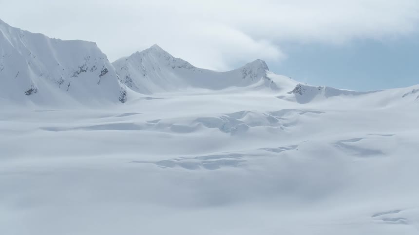 Snowy mountains tower over the nearby snow covered plains and hills on a cloudy day