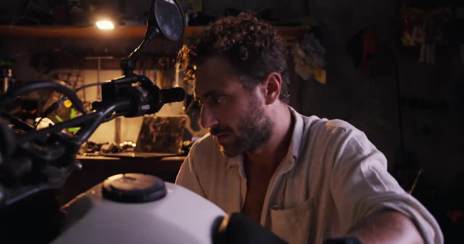 A person works on a motorcycle in a dimly lit garage with tools in the background