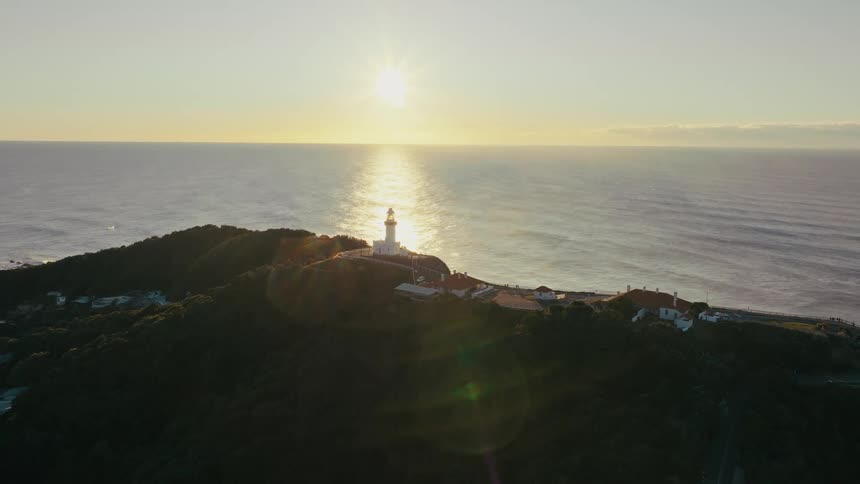 Aerial view of an island with some houses and a lighthouse by the sea with the sun on the horizon