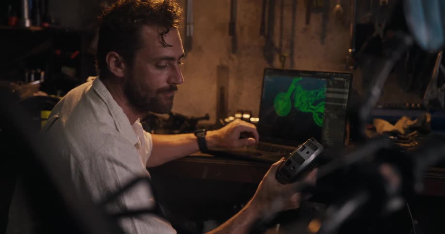 Man holds a mechanical part while designing a motorcycle on his computer