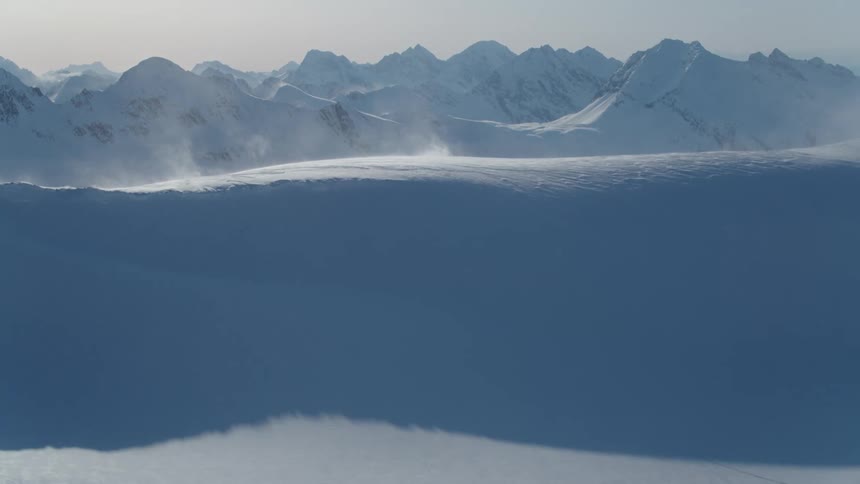 A windy day in the mountains with snow blowing across a snowy landscape
