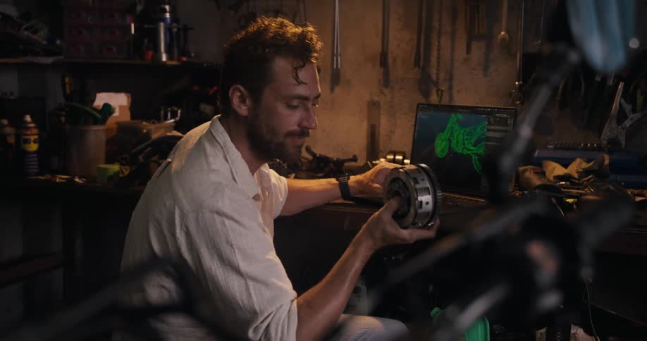 A person at a workbench examines a mechanical part while looking at a laptop screen