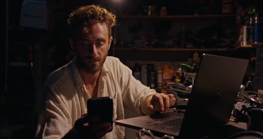 Man sitting in a workshop uses both a smartphone and a laptop while working