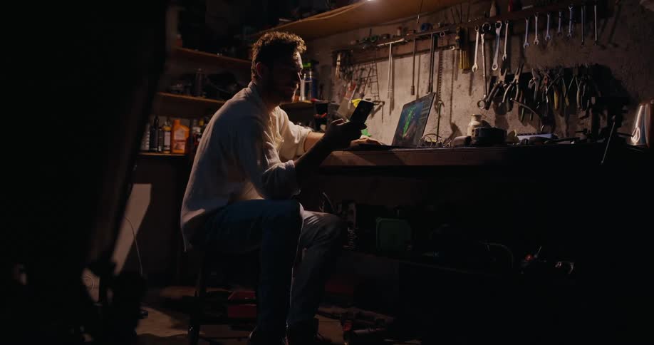 A man at a workbench in a dimly lit workshop holding a smartphone and looking at a laptop screen