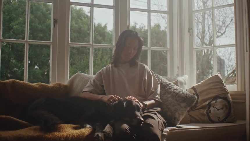 A woman sits by a window petting a dog while sunlight streams in