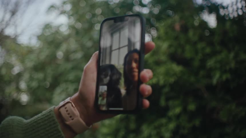 A person holds a smartphone having a video call with someone on the other side of the screen with a blurred background of lush greenery