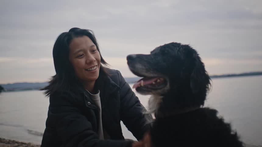 A woman smiles while petting a large black dog by a lake