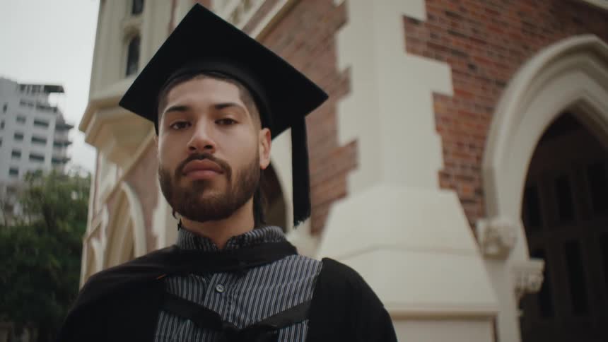 Young male with facial hair stands outside near brick building wearing graduation cap