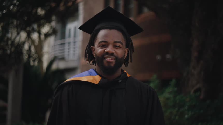 A young man wearing a graduation cap and gown smiles at the camera