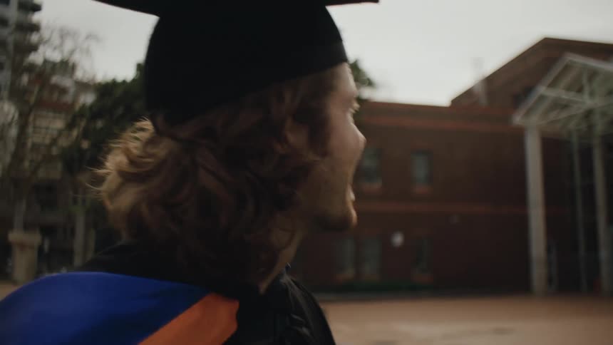A young man in graduation attire including a cap and gown stands outdoors near a modern building