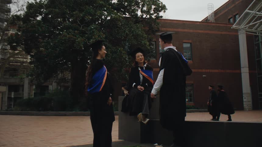 Three people in graduation gowns and caps stand and sit on a low wall outside a building