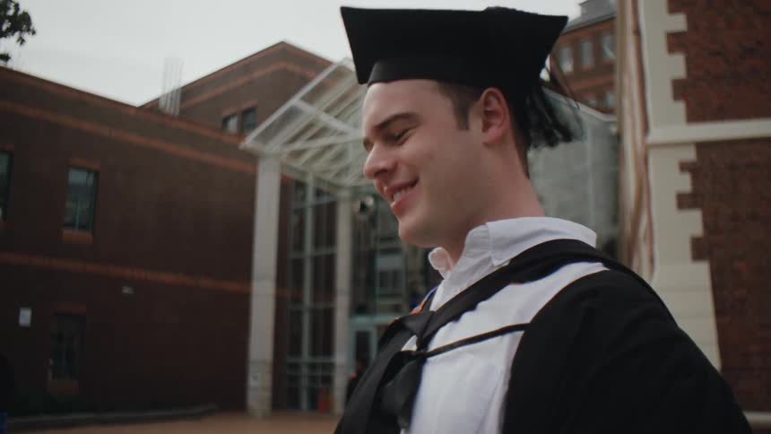 A graduate in cap and gown stands a brick building