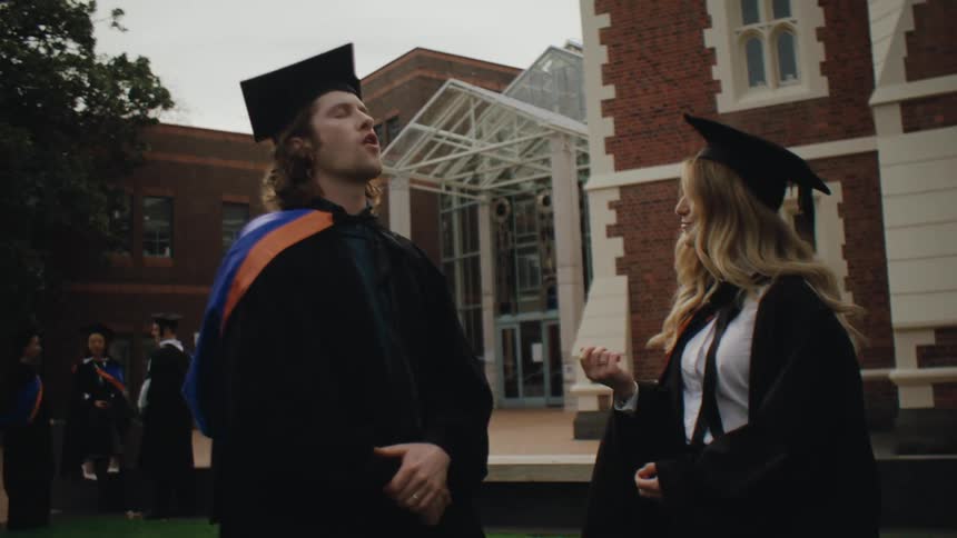 Two graduating students stand outside a school building and talk