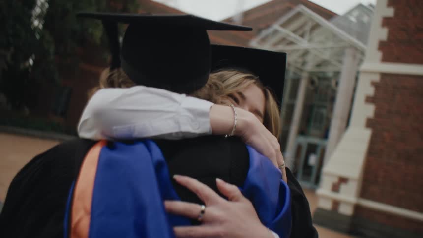 A couple of friends in graduation gowns and caps celebrate by hugging each other