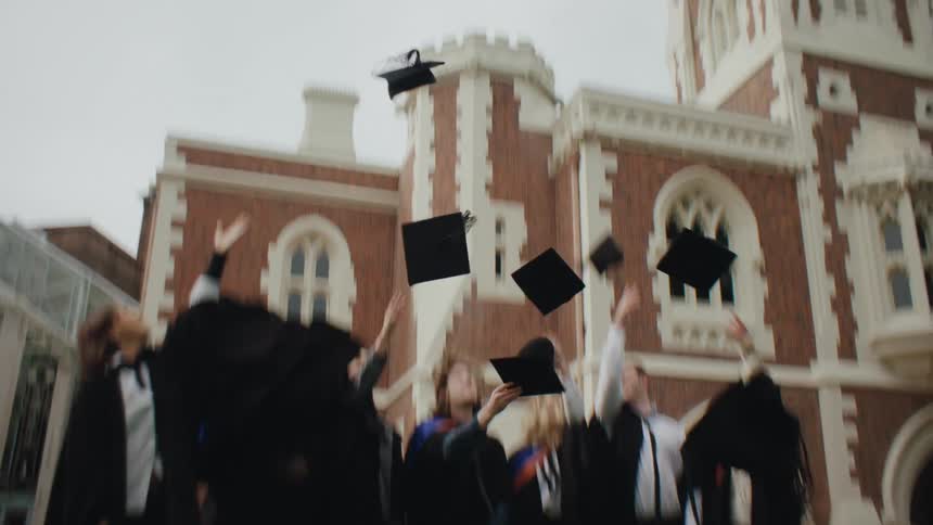 A group of graduates dressed in caps and gowns celebrate outside a historic brick building tossing their caps into the air with joyful expressions