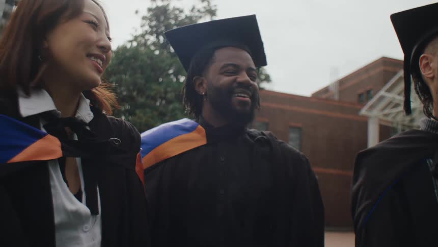 Men and women wearing graduation cap and gown smile as they stand outdoors