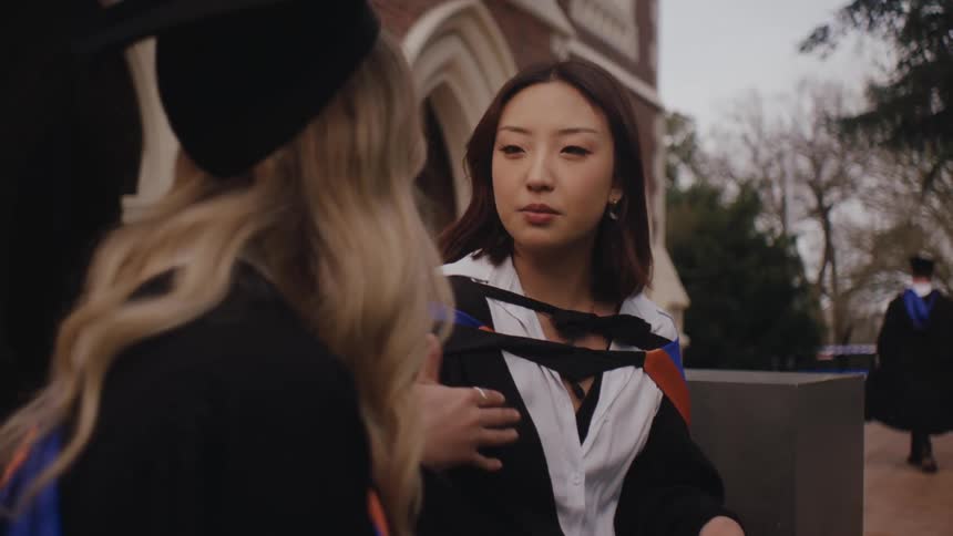 Two graduates in academic robes and caps talk to each other in front of brick building