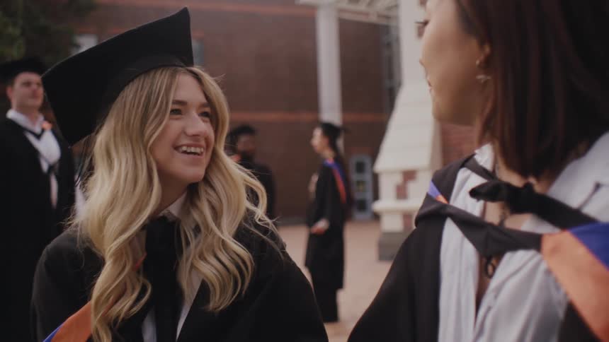 Two women in graduation gowns are laughing and talking to each other outdoors