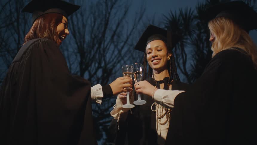 Three women toasting glasses of champagne while wearing graduation gowns