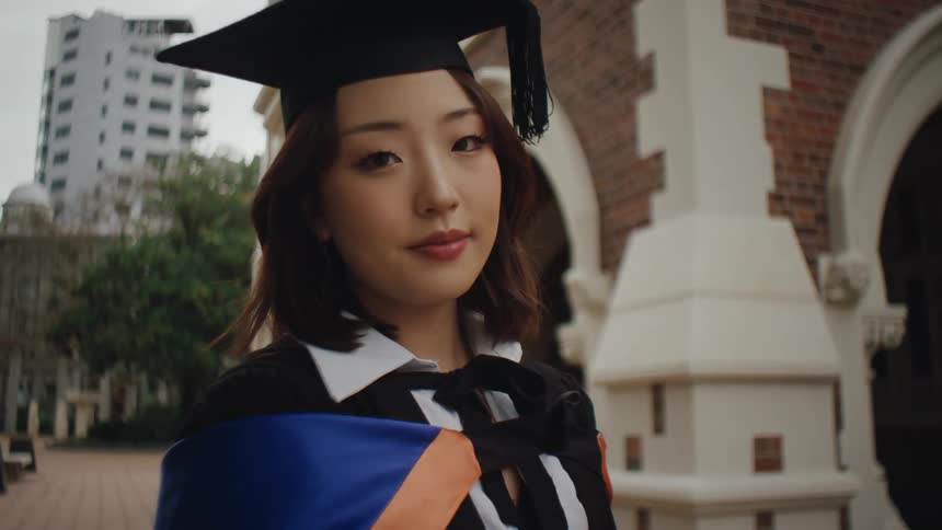A young asian woman in a graduation outfit poses for a photograph outdoors in front of a building