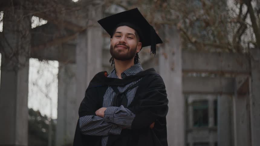 A graduate student stands with crossed arms wearing a cap and gown