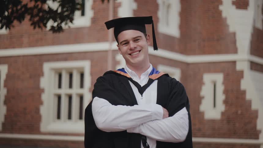 A graduating student in cap and gown smiles outside of a school building