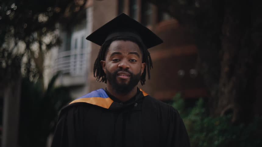A young man wearing a graduation cap and gown smiles at the camera