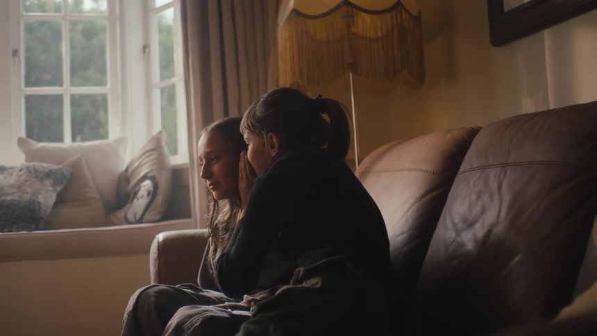 Two girls sit on a couch one whispering something in the others ear