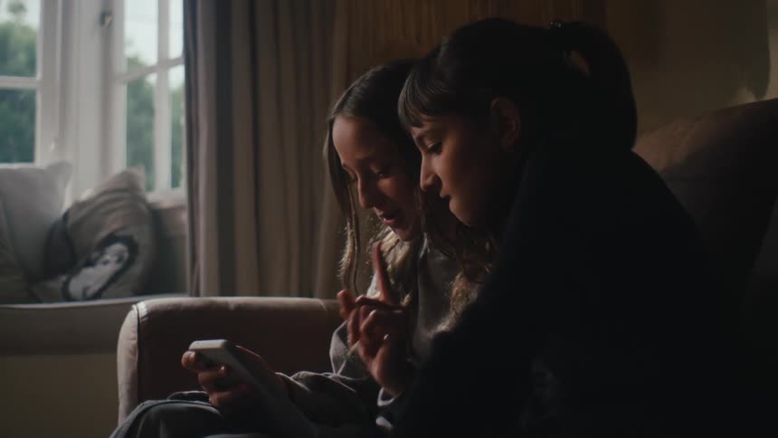 Two little girls sitting on the sofa in the living room and interacting with a smartphone