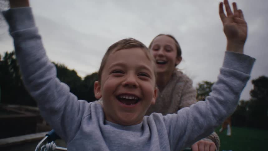 A girl pushes a boy in a shopping cart through a park