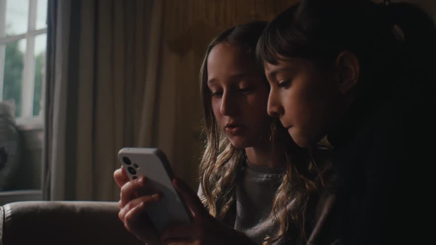 Two girls sitting in the living room talk while sharing a smartphone