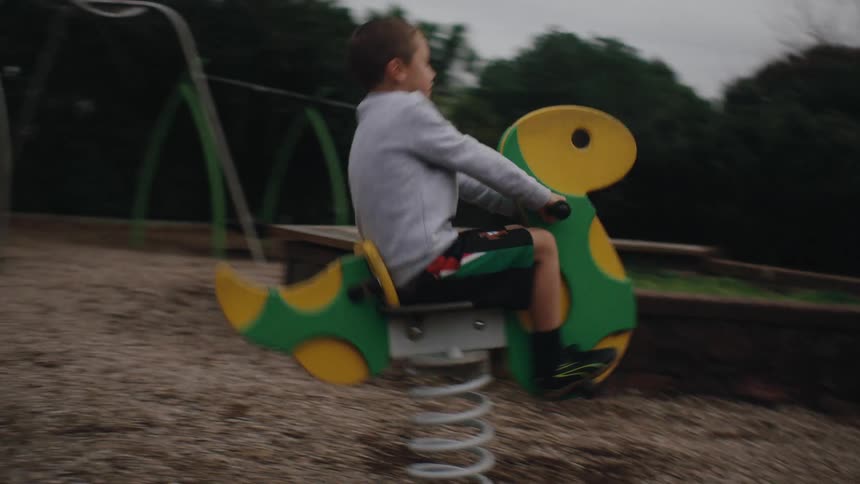 A young boy rocks back and forth on a colorful springmounted playground toy his focus steady as the overcast sky and surrounding greenery create a calm quiet atmosphere in the park