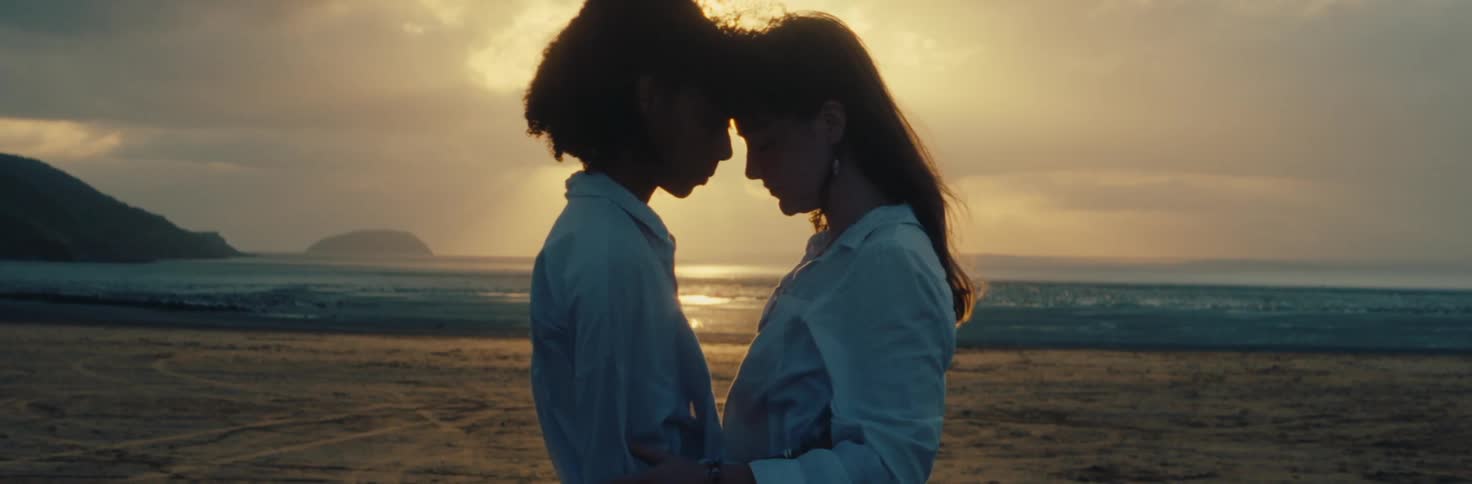 Two women stand nose to nose backlit by sunlight on the beach