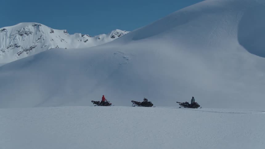 Three people stand next to snowmobiles on a snowy mountainous landscape