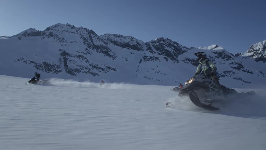 People ride snowmobiles in a snowy landscape with mountains in the background