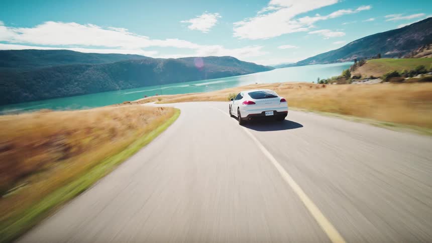 A white sports car drives along a winding road overlooking a lake