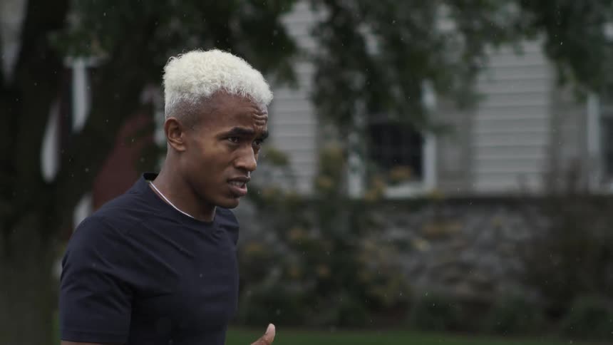 A person with short bleached blonde hair jogs through the neighborhood in light rain