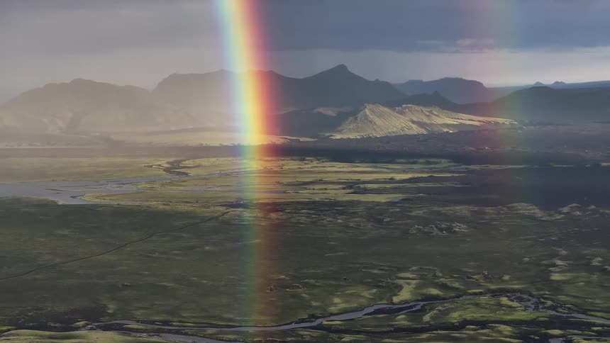 Vast landscape with mountains in the background and a rainbow descending from a cloudy sky to the green valley below