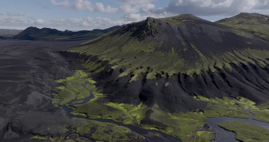 The stark contrast of black volcanic ash and lush green moss creates a dramatic landscape where winding rivers carve their way through the rugged terrain