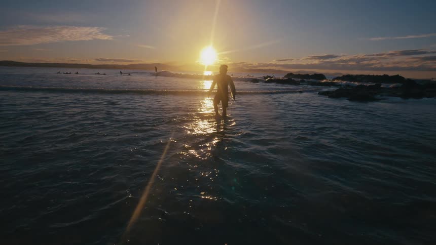 Silhouette of a person with a surfboard walking through shallow water towards the sunset