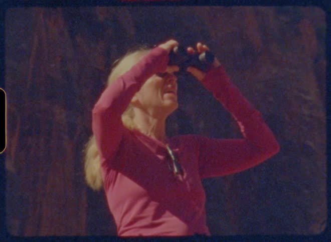A woman stands in front of towering red cliffs lifting binoculars to her eyes to take in the stunning rugged landscape around her
