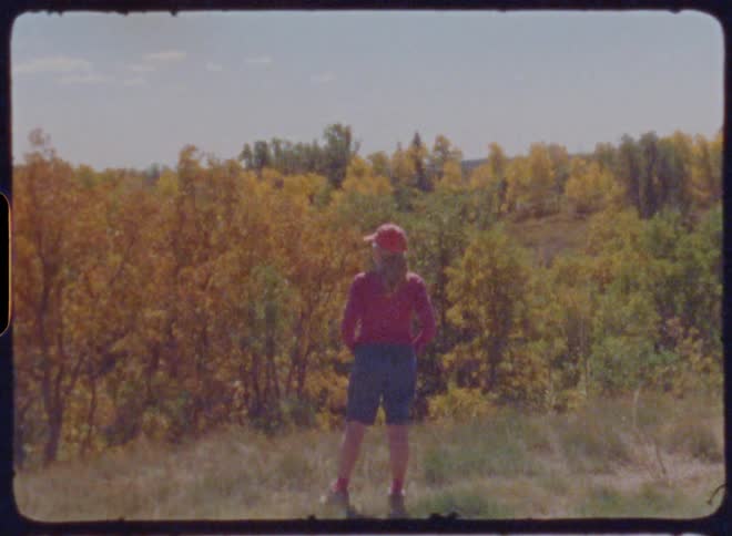 A woman stands on the side of a road looking at a forest with autumn colored trees