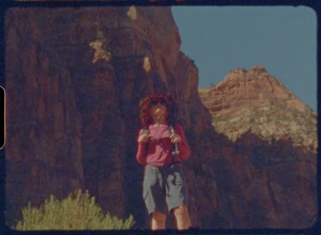 Woman is standing at the base of a rocky cliff with clear sky above and some vegetation in the background