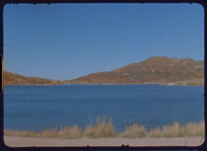 A calm lake surrounded by rolling hills under a clear blue sky captured on vintage film