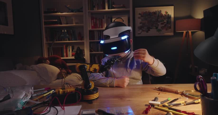 Boy sitting at a messy table wearing a virtual reality headset and a mechanical arm on an electronic device