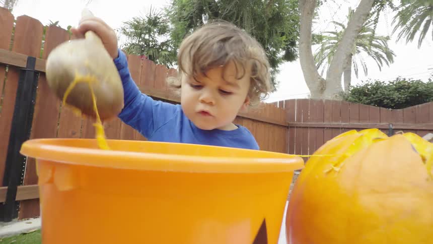 Little boy holding a wooden spoon and scooping pieces of pumpkin into a bucket nearby